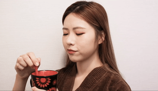 穿著棕色上衣、在樸素背景下閉著眼睛攪拌紅黑杯子的人。.